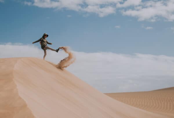 Persoană aflată pe o dună de nisip, sub un cer albastru, lovind cu un picior un jet de nisip.