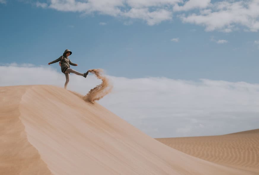 Persoană aflată pe o dună de nisip, sub un cer albastru, lovind cu un picior un jet de nisip.