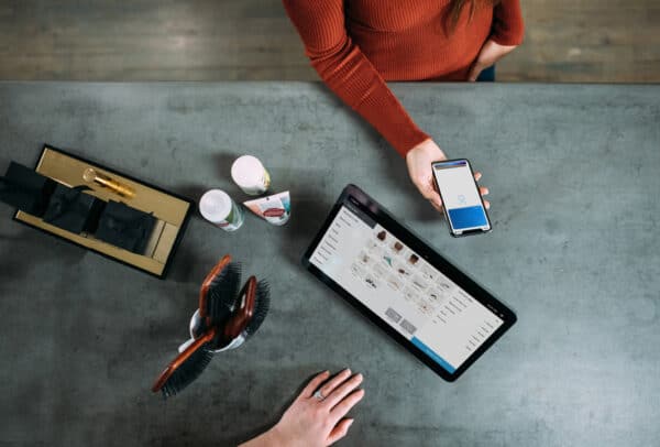 Două persoane la un blat, una ținând un smartphone cu un ecran de plată, cealaltă lângă o tabletă care afișează o listă de produse; perii de păr și articole mici sunt, de asemenea, pe tejghea.