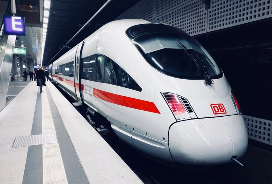 Un tren modern de mare viteză Deutsche Bahn de culoare albă, cu bandă roșie, este oprit pe o platformă interioară a unei gări.