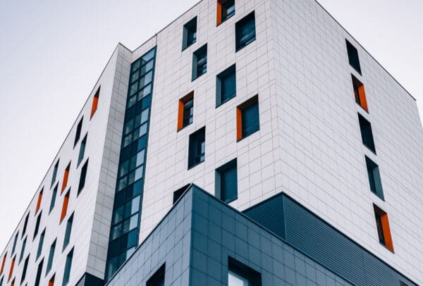 Clădire înaltă modernă cu o grilă de panouri albe și albastre, cu ferestre dreptunghiulare, unele cu accente portocalii, fotografiată dintr-un unghi scăzut pe un cer senin.