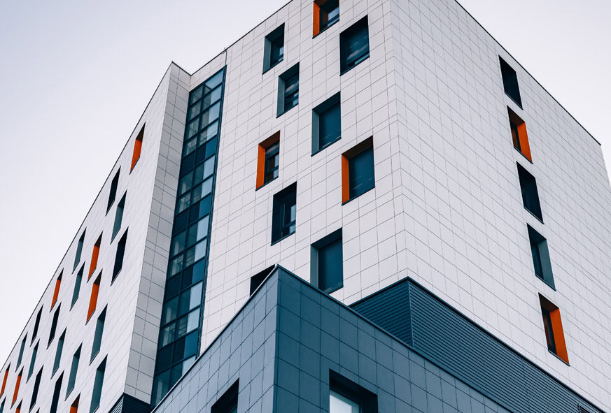 Clădire înaltă modernă cu o grilă de panouri albe și albastre, cu ferestre dreptunghiulare, unele cu accente portocalii, fotografiată dintr-un unghi scăzut pe un cer senin.