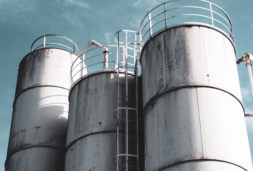 Trei rezervoare mari, cilindrice, metalice, cu scări și balustrade, se află pe un cer albastru.