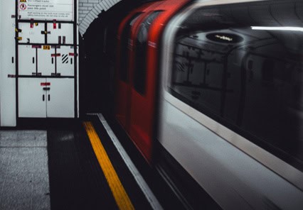 Un tren de metrou roșu și alb intră într-o stație subterană, cu panouri de control și semne de avertizare vizibile pe perete.