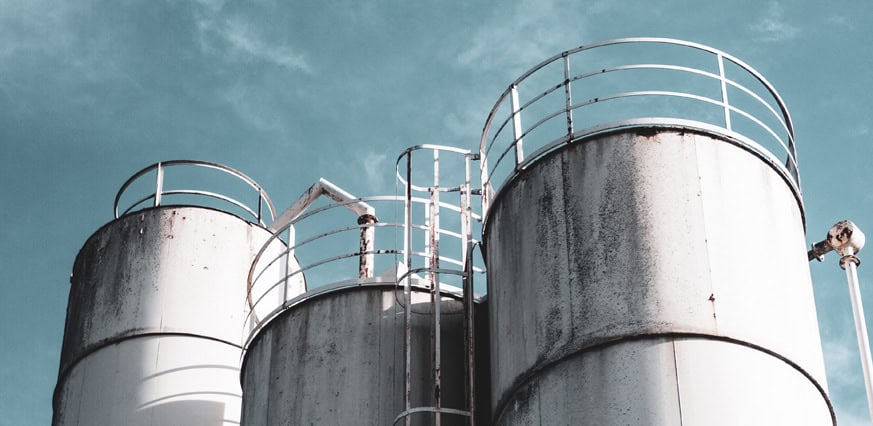 Trei rezervoare industriale înalte, albe, cu balustrade metalice sunt prezentate pe un cer albastru. Rezervoarele prezintă urme vizibile de rugină și intemperii.