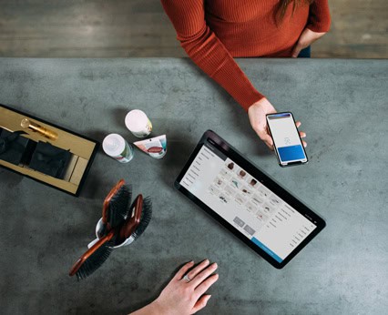 O persoană ține un smartphone care afișează o aplicație de plată fără contact în apropierea unei case de marcat cu tabletă, în timp ce o altă mână operează casa de marcat pe un blat cu diverse articole.